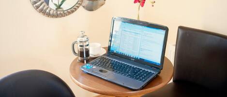 A laptop on a wooden table with a coffee pot and a cup, a mirror with a flower, and a chair.