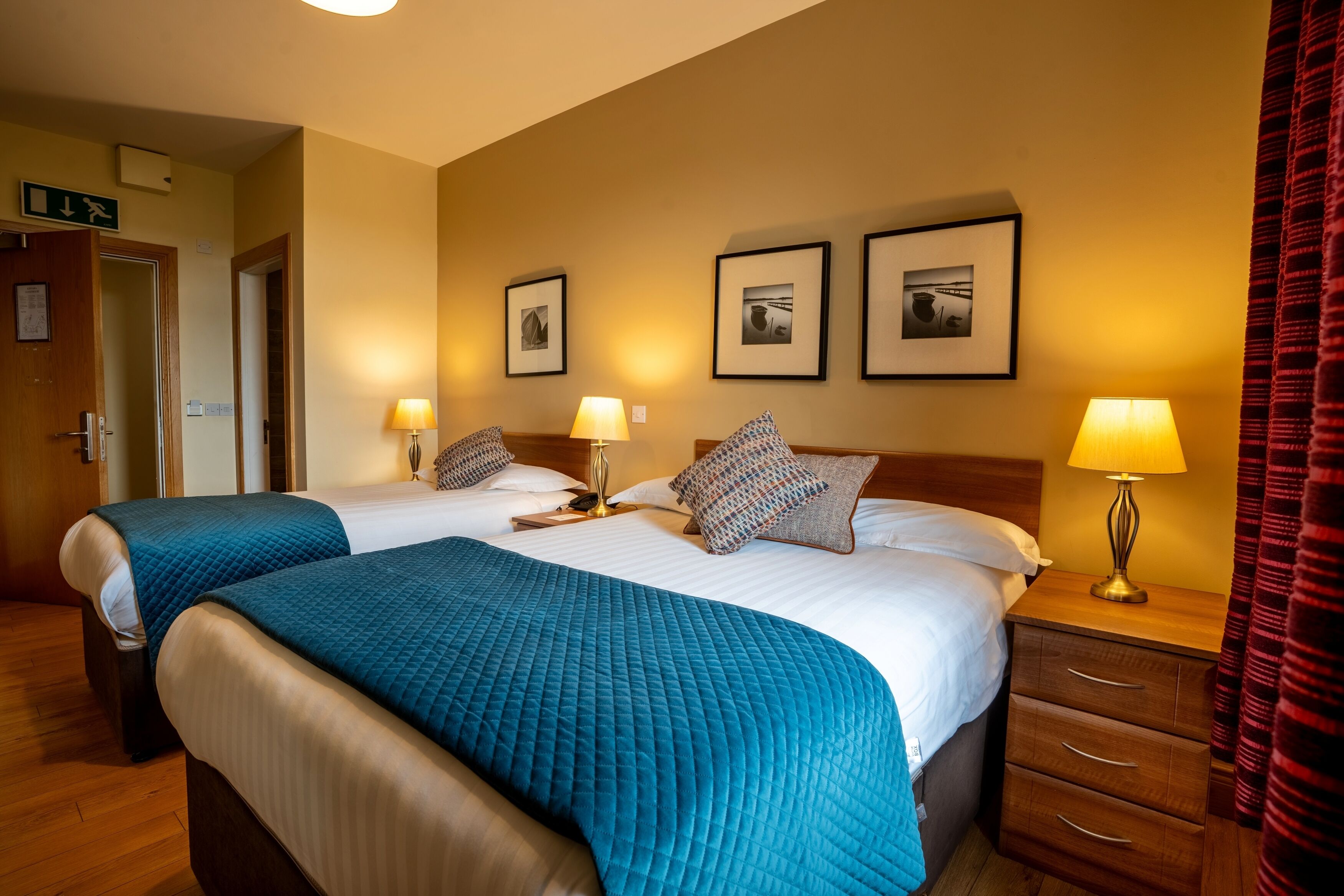 A hotel room with two beds, each with a blue quilted bedspread and white linens. There are two bedside lamps, a wooden nightstand with drawers, and framed pictures on the wall.