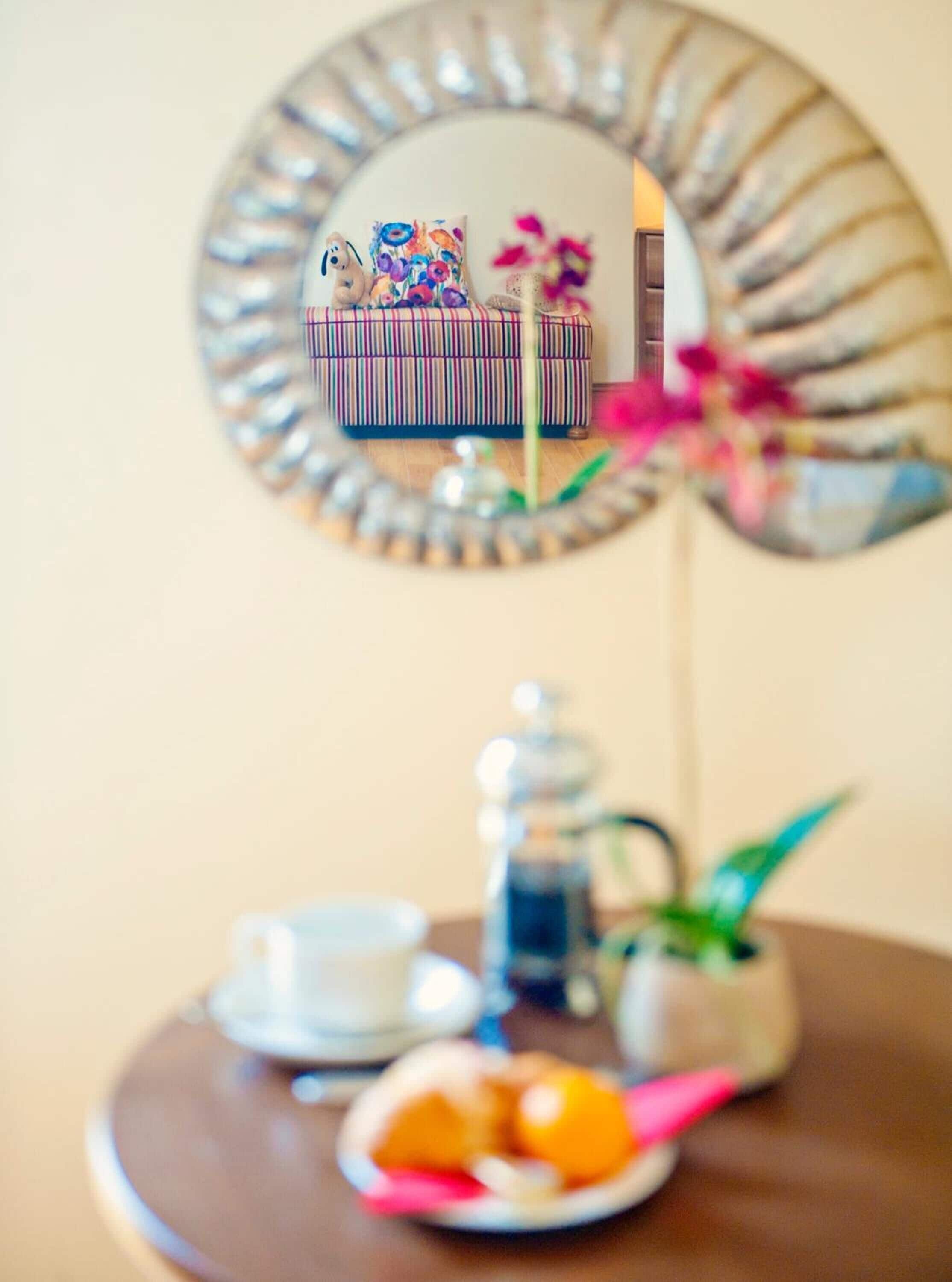 A round mirror with a decorative frame reflecting a striped sofa and a small table with a teapot, cups, and a plate of fruit.