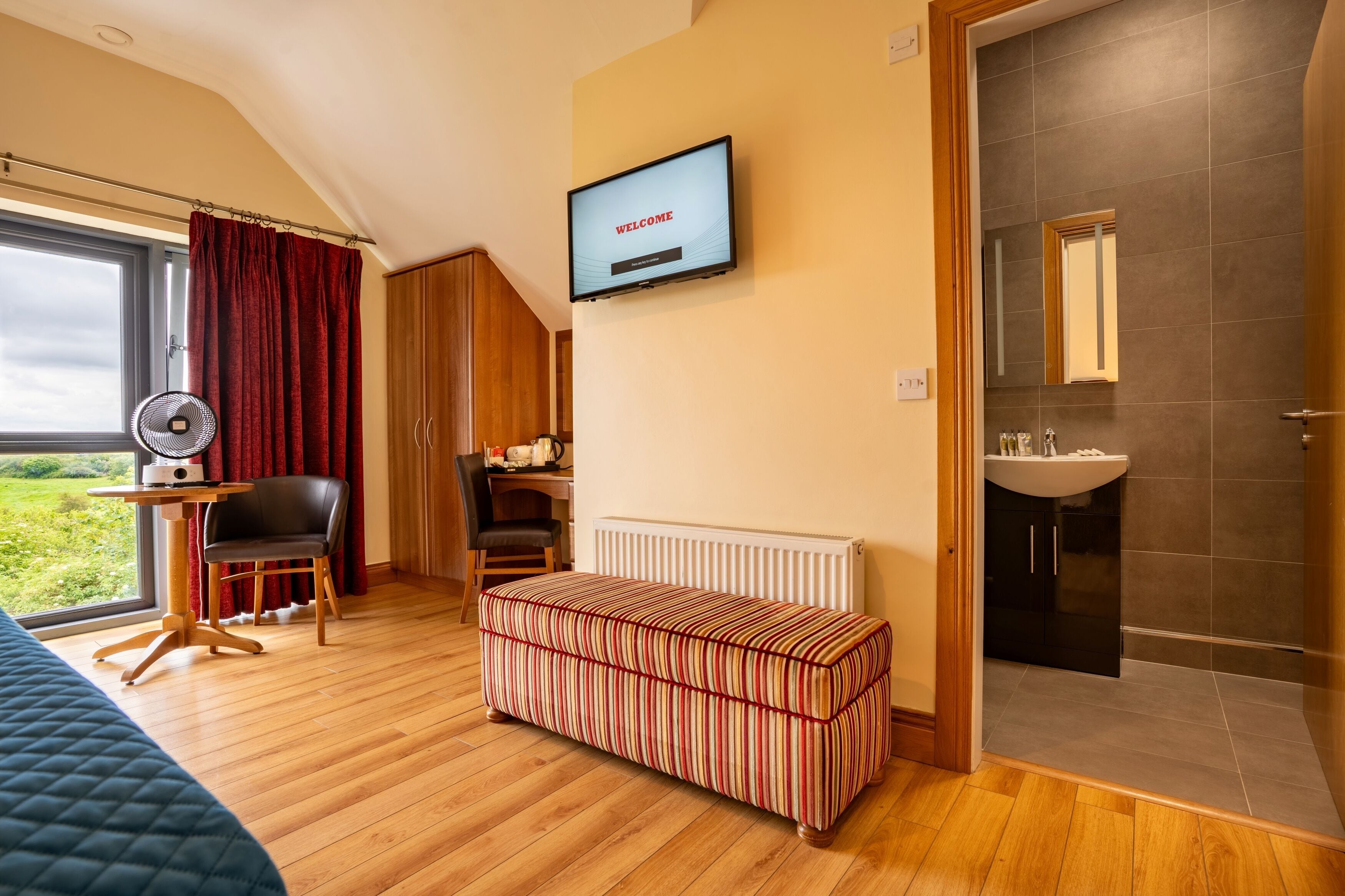 A hotel room with a bed, a desk with a chair, a TV displaying "WELCOME", a window with curtains, a radiator, a bench, and a bathroom with a sink and mirror.
