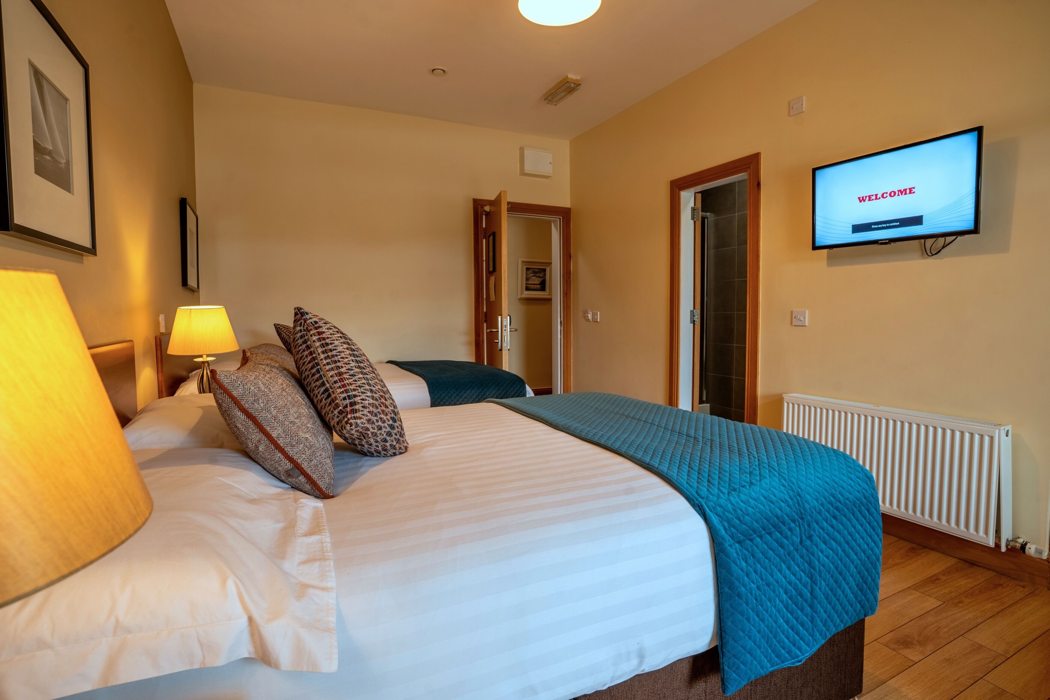 A hotel room with a bed, a television displaying "WELCOME," a lamp, and a door leading to another room.
