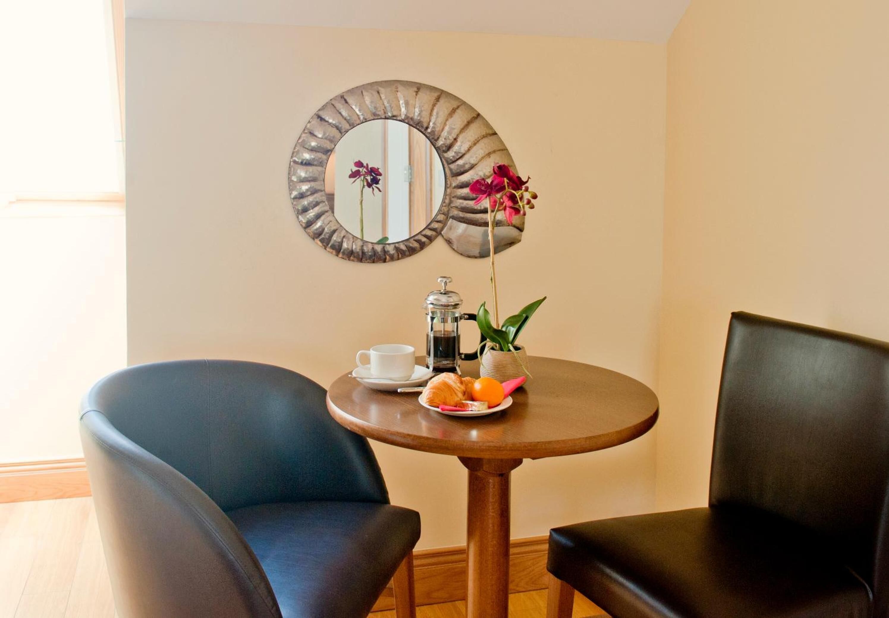 Una habitación con dos sillones de cuero negro, una pequeña mesa redonda de madera con una taza blanca, una cafetera de plata, un plato con comida y una planta en maceta con flores.