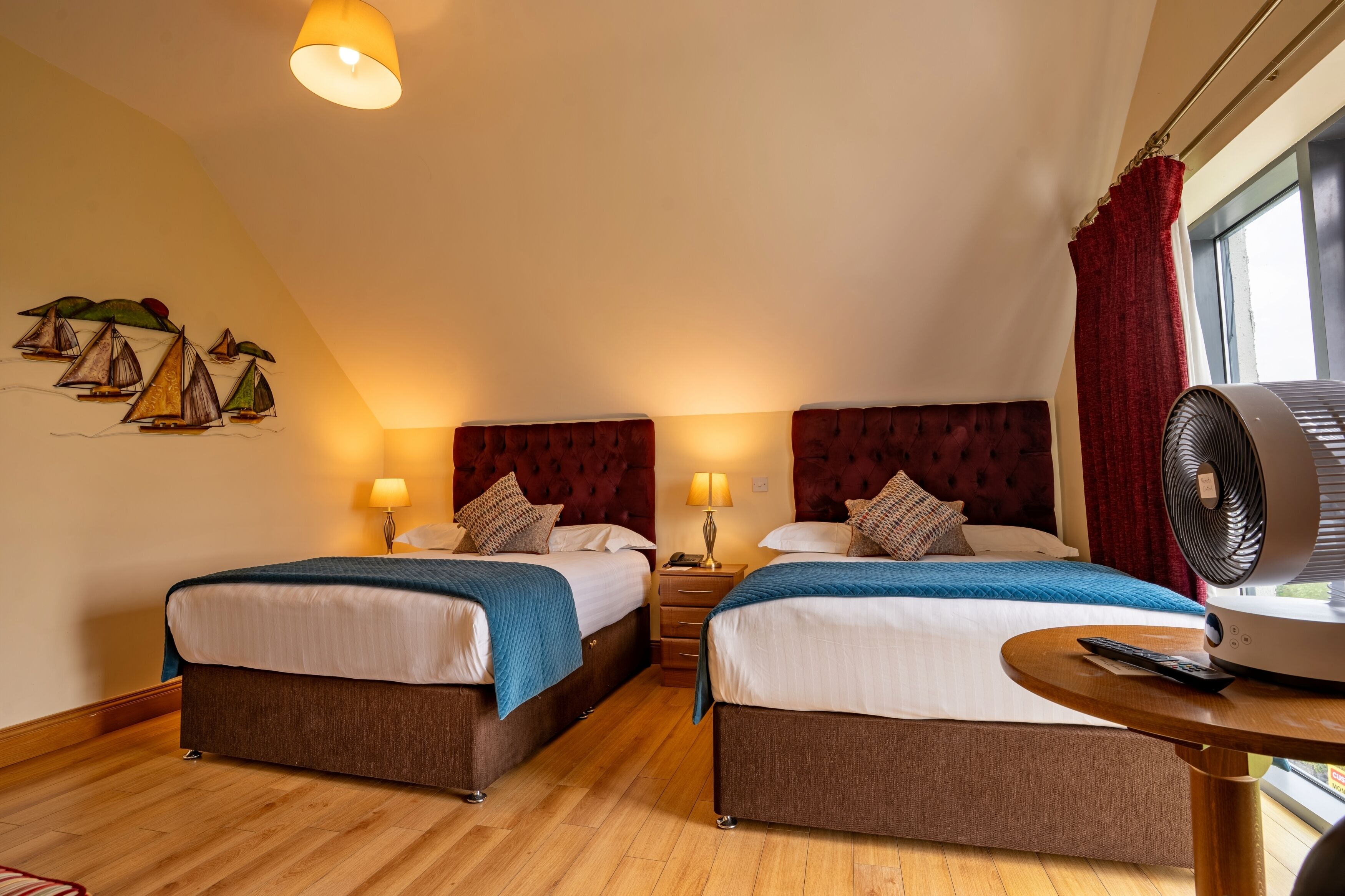 A hotel room with two beds, a wooden floor, a ceiling fan, and a window with red curtains.