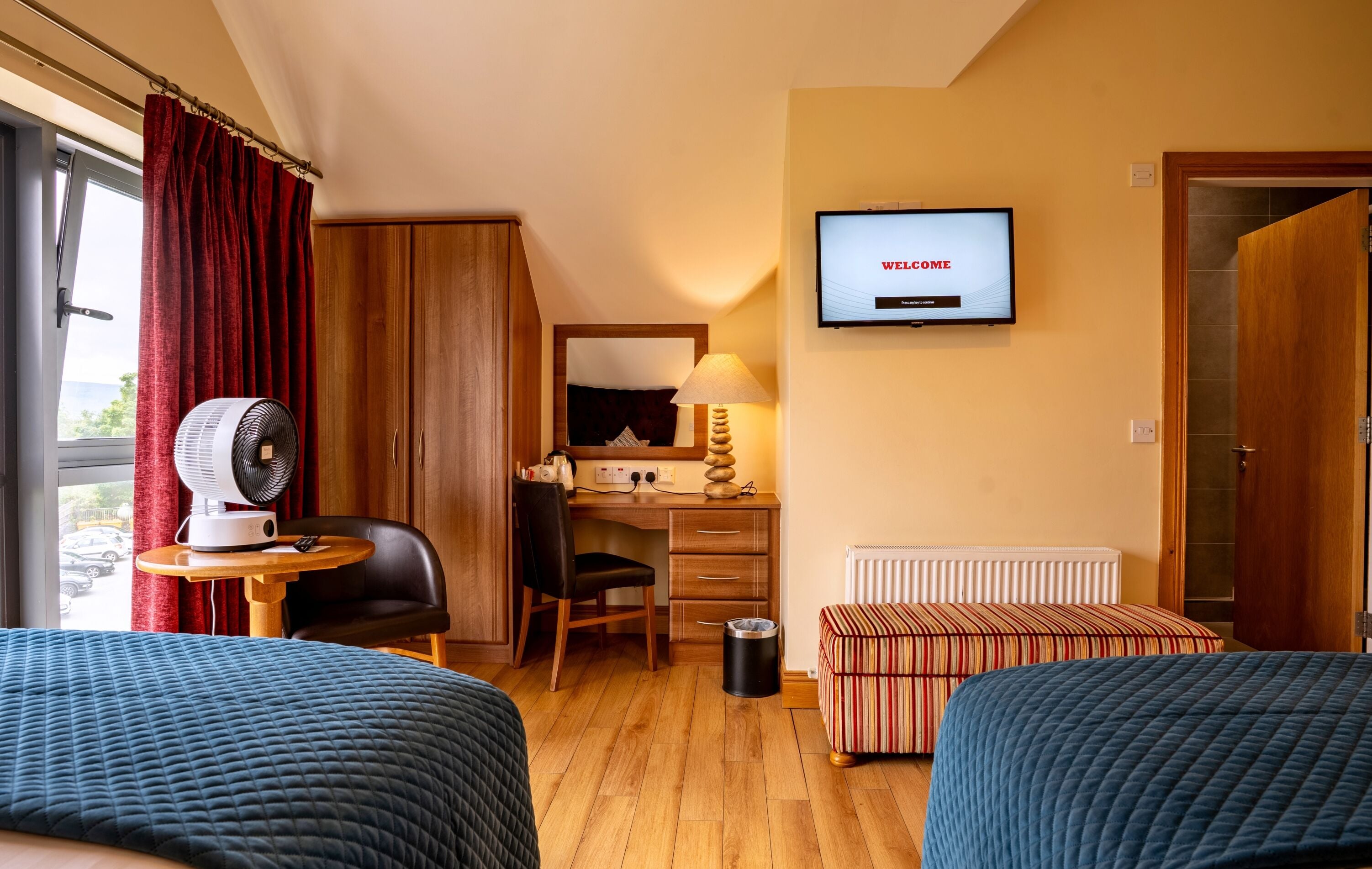 A hotel room with two beds, a desk with a computer, a TV displaying "WELCOME", a window with curtains, and a radiator.