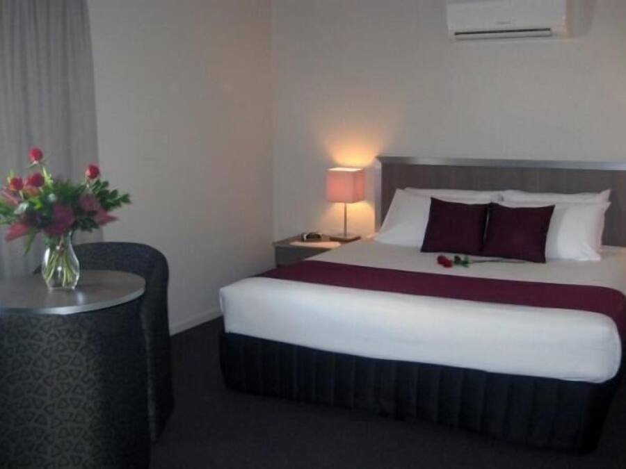 Ein Hotelzimmer mit Bett, Nachttisch mit Lampe, Blumen in einer Vase und einer Klimaanlage an der Wand.