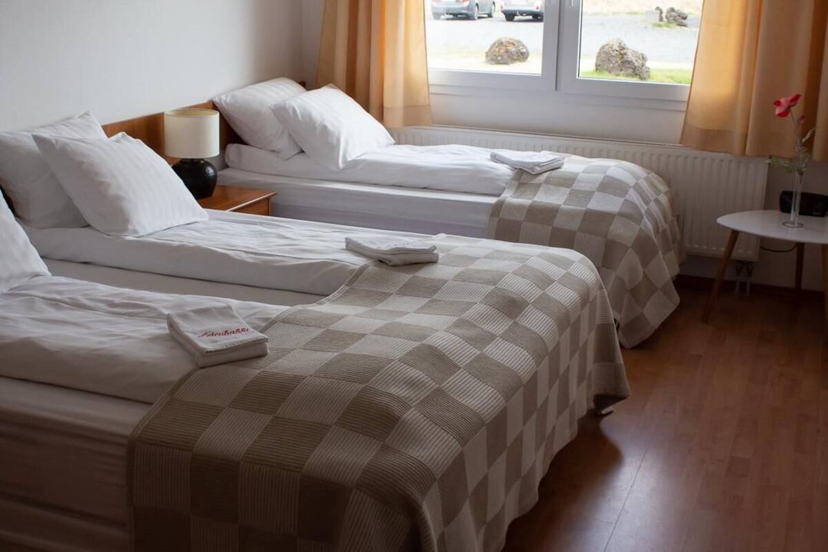 A hotel room with two beds, a small table, a lamp, and a view of the outside.