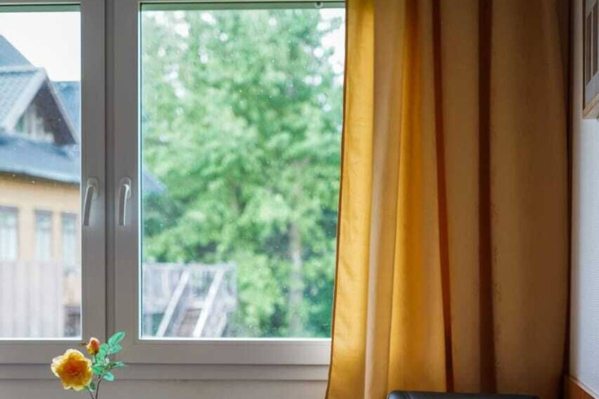 A hotel room with a bed, a chair, a window with curtains, and a vase with a flower.