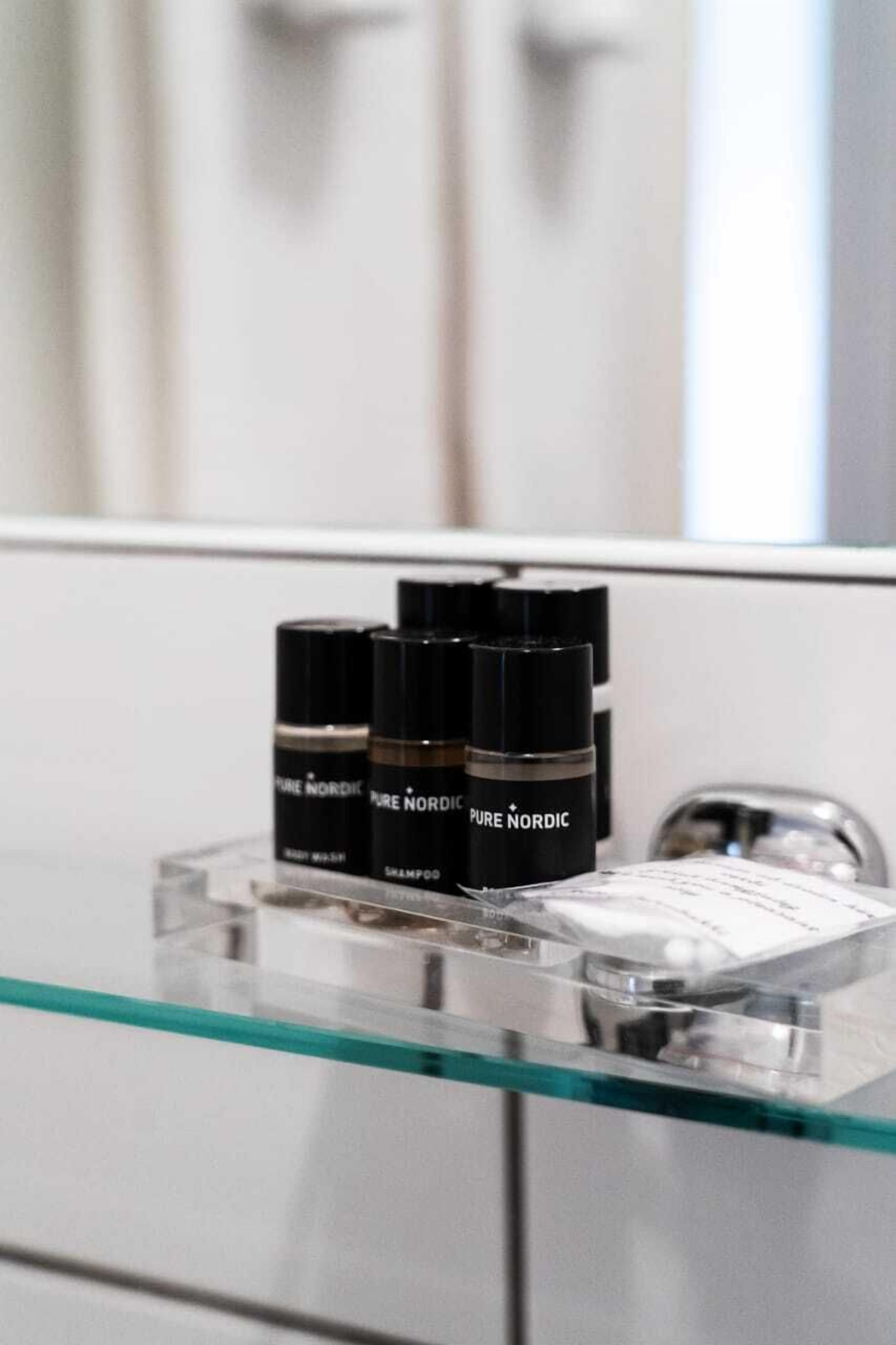 Une salle de bain avec une étagère en verre sur laquelle sont posées trois bouteilles noires portant l’étiquette « PURE NORDIC » et un récipient blanc.