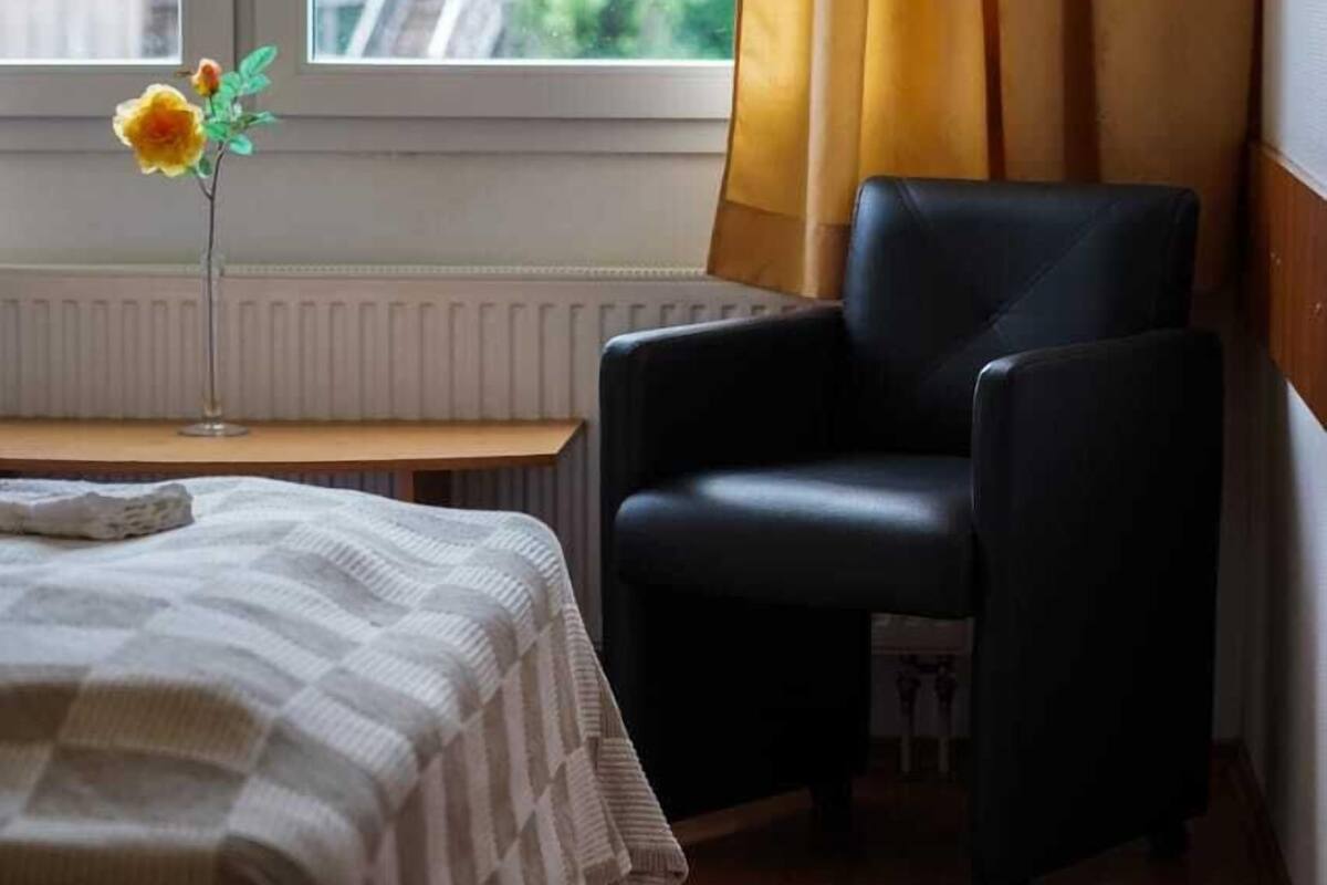 A hotel room with a bed, a chair, a window with curtains, and a vase with a flower.