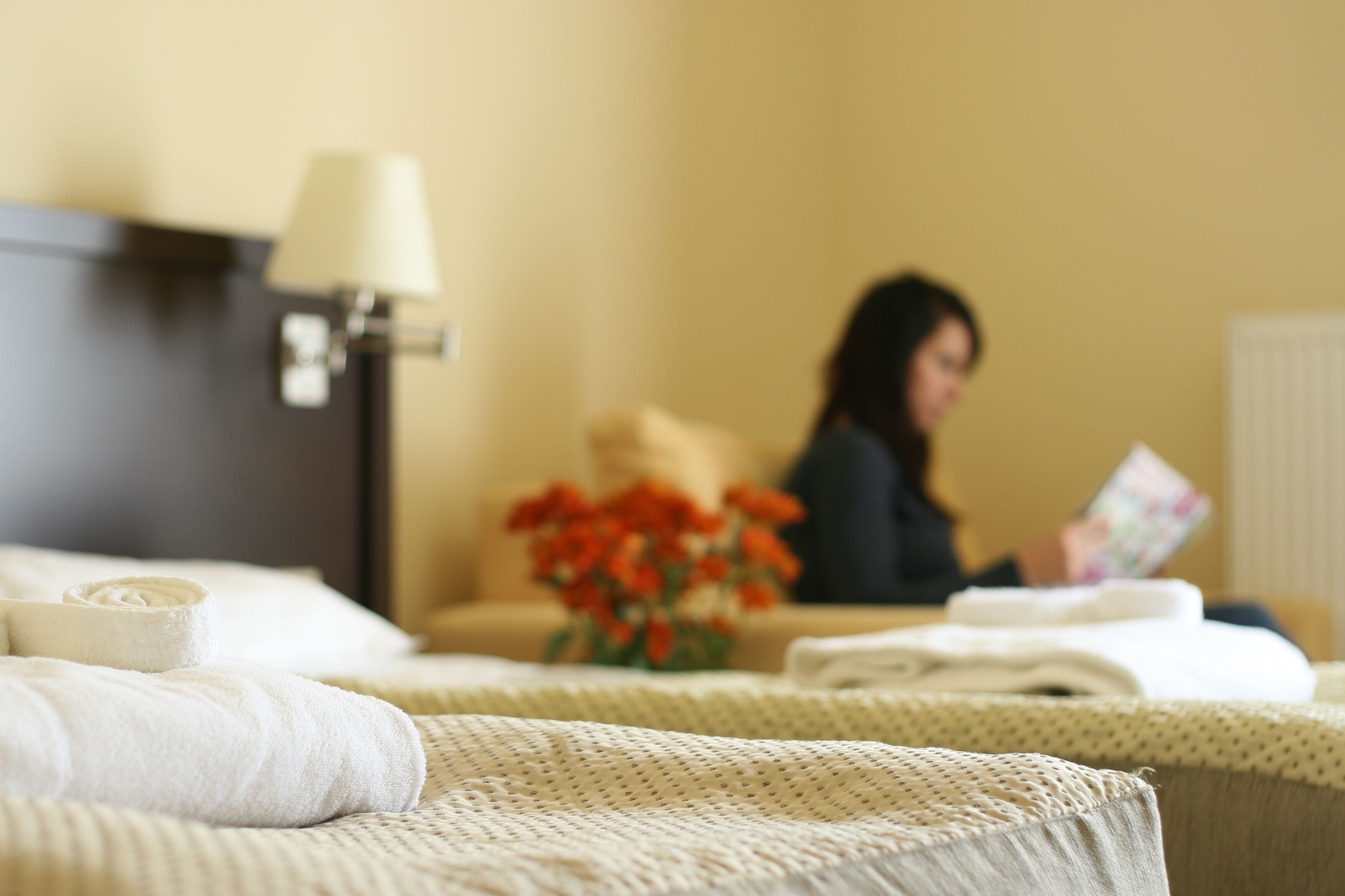 Una cama bien hecha con una toalla enrollada, una lámpara y un florero con flores sobre una mesita de noche.