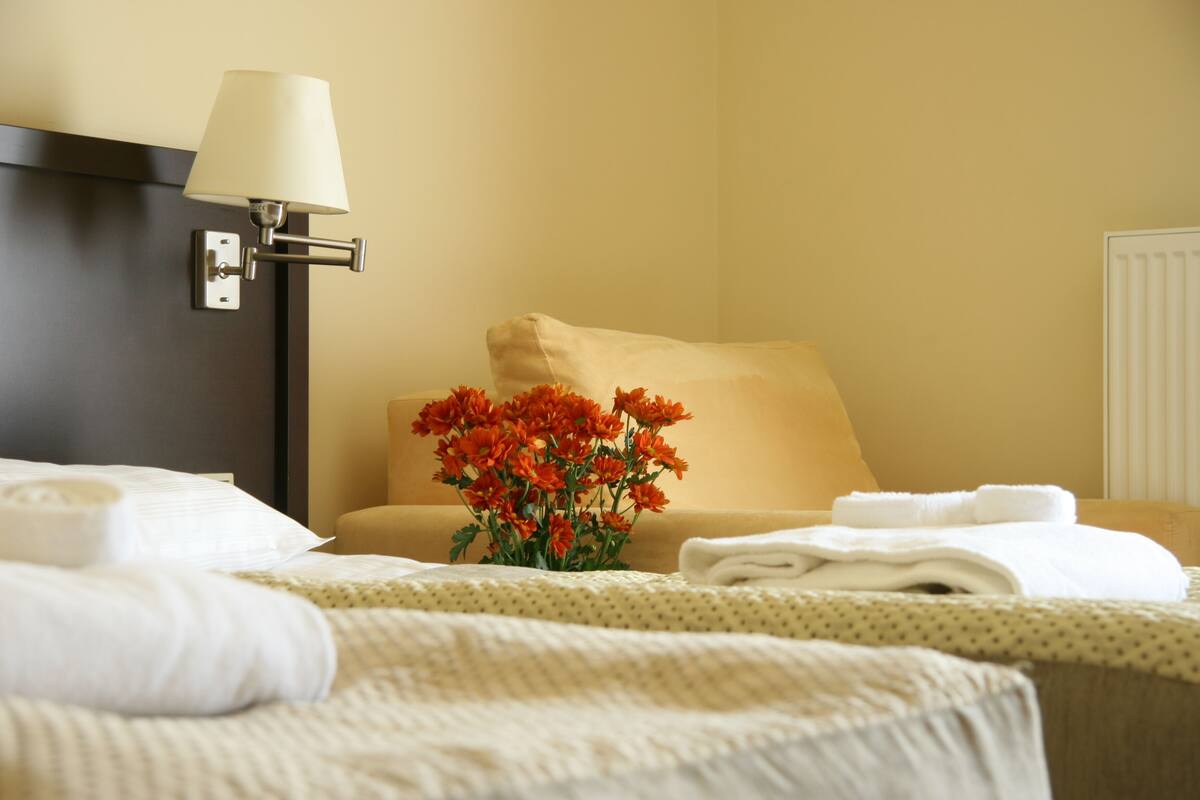 Habitación de hotel con cama, mesita de noche, lámpara, un jarrón con flores y un radiador.