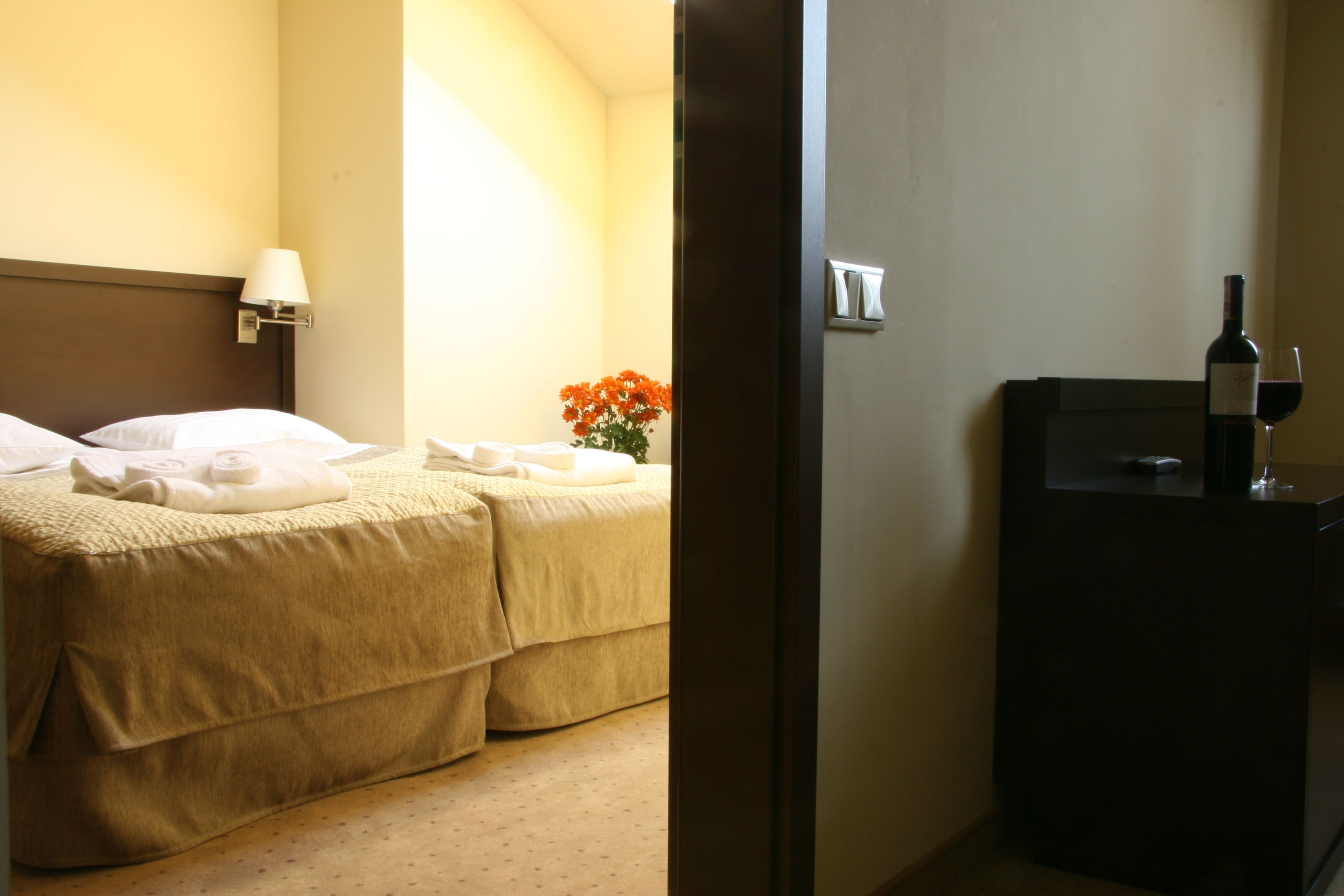 A hotel room with two beds, a nightstand with a lamp, a vase with flowers, a minibar, and a wine bottle.