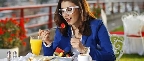 A person dining outdoors with a table set for breakfast, including a fruit bowl, coffee, and a glass of orange juice.