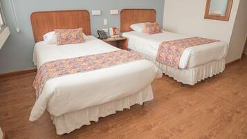 A hotel room with two beds, each with a floral bedspread and white bedding. There is a bedside table with a phone and a small lamp. The room has wooden flooring and light blue walls.