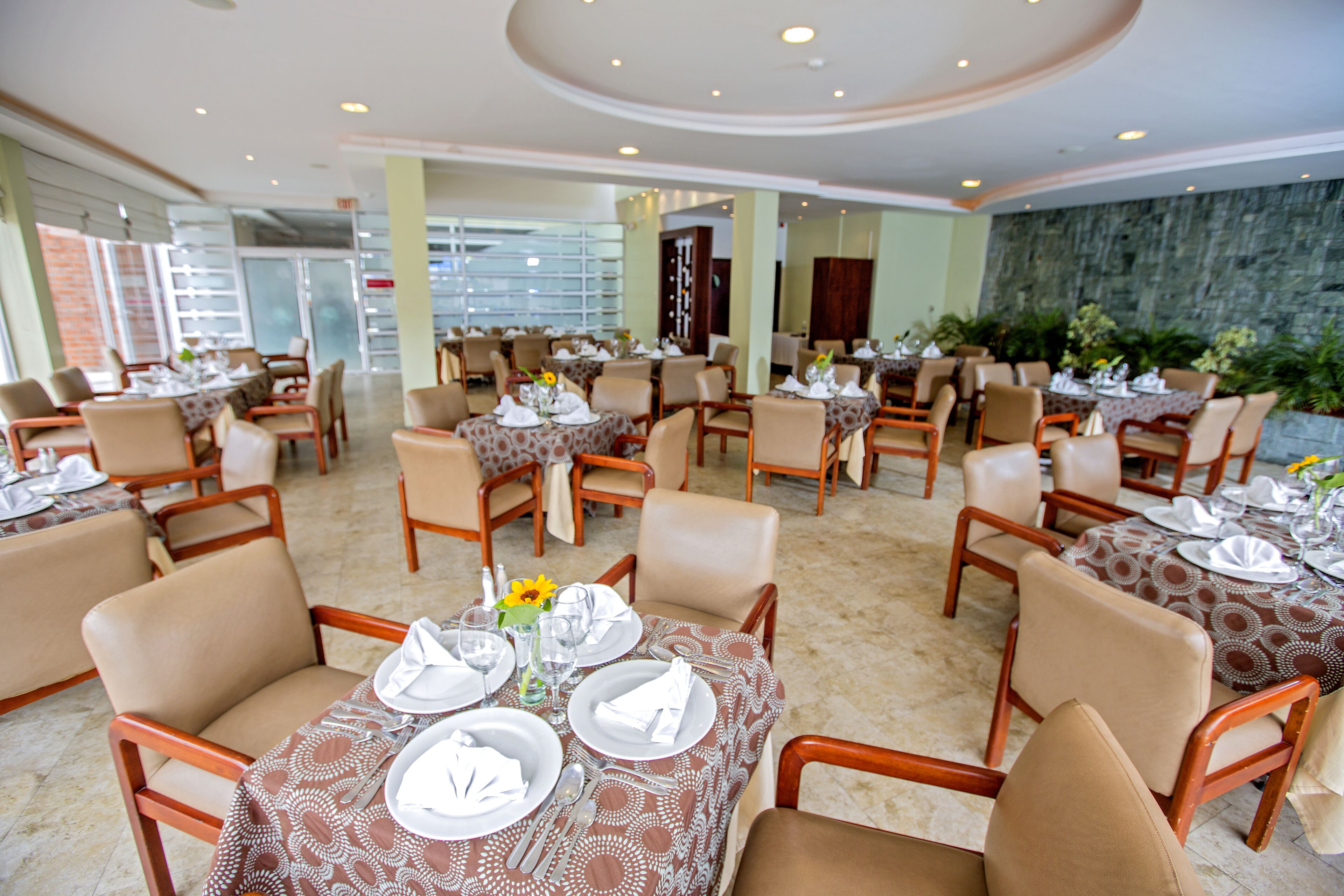 Un restaurant avec plusieurs tables dressées pour le service, agrémenté d’une nappe à motifs et de chaises de couleur beige.