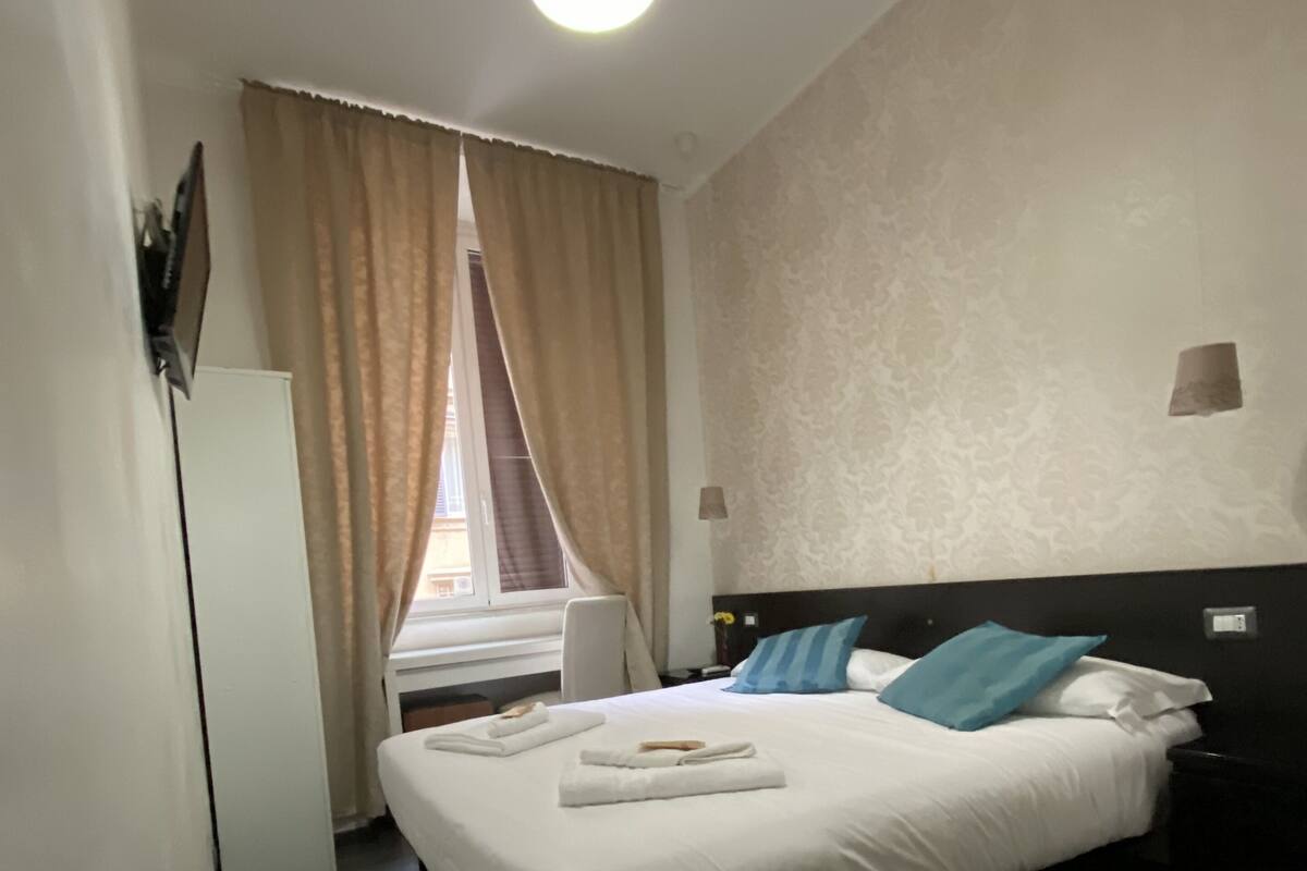 A hotel room with a bed, a desk, a chair, a window with curtains, and a ceiling light.