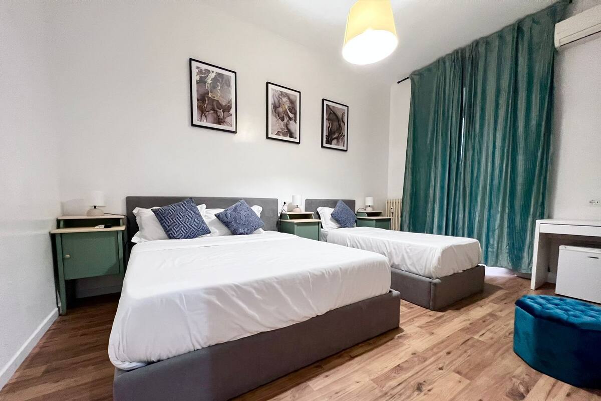 A hotel room with two single beds, a desk, a chair, a small table, and a green curtain.