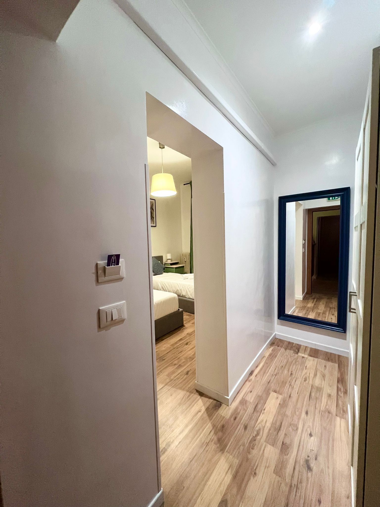 A hallway with a wooden floor, a bed with white bedding, a bedside table with a lamp, and a door with a mirror.