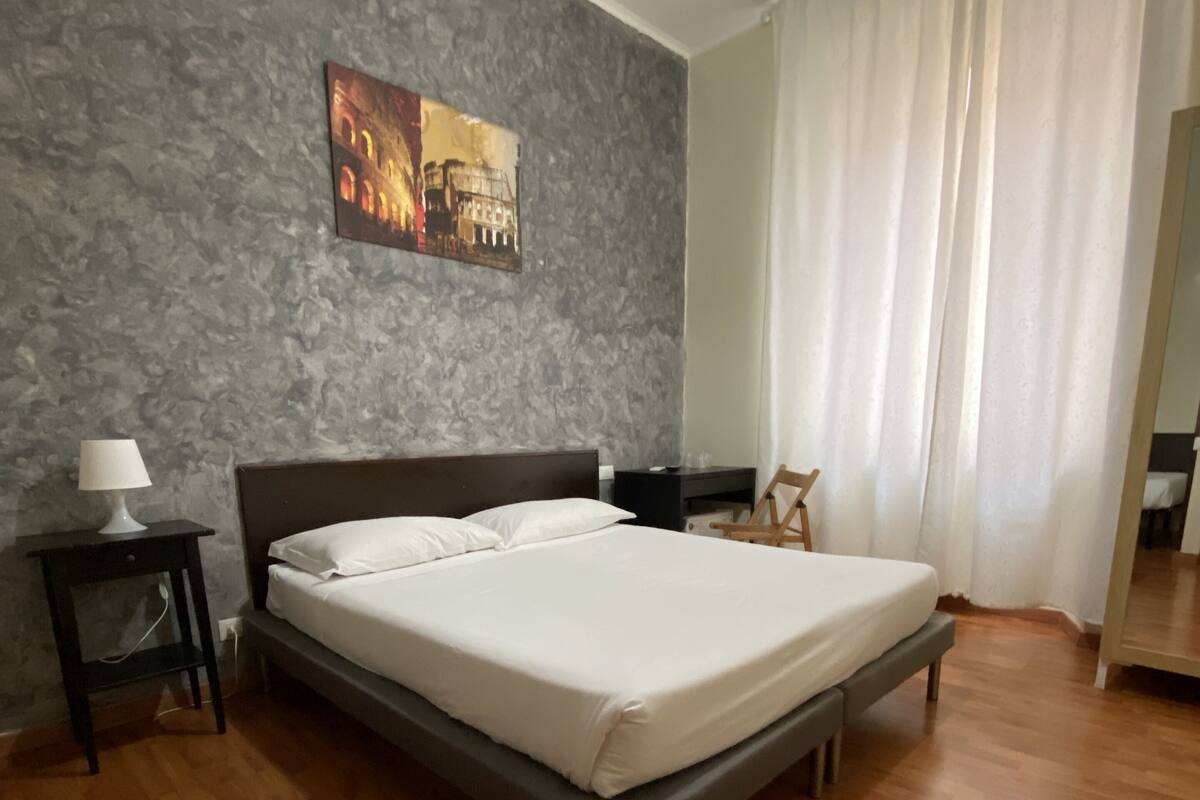 A bedroom with a bed, a nightstand, a lamp, a painting on the wall, and a window with curtains.
