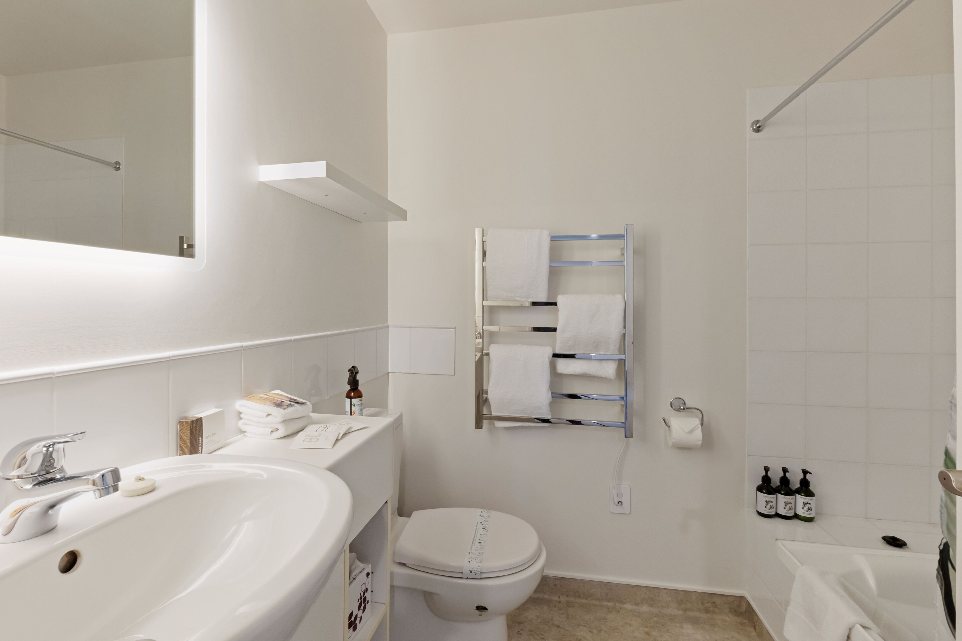 Un baño moderno con lavamanos, inodoro y bañera blancos. Hay un perchero con varias toallas y un dispensador de jabón en la pared.