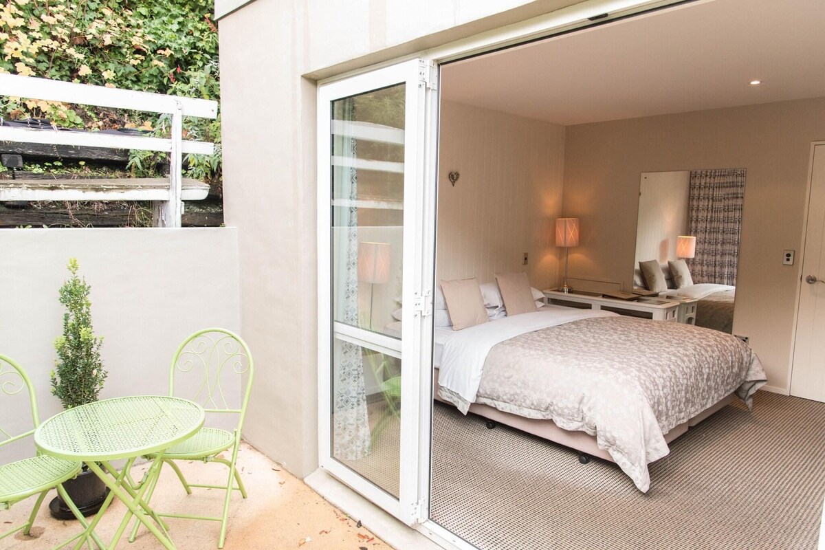 A bedroom with a bed, a desk, and a chair. There is a small table and a potted plant outside the room.