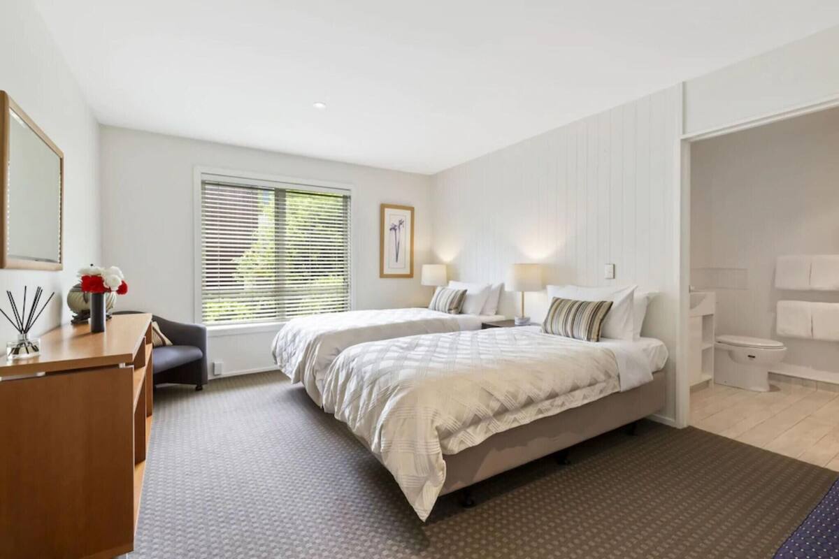 Ett hotellrum med två sängar, ett skrivbord, en stol, ett fönster med persienner, ett badrum och en spegel.