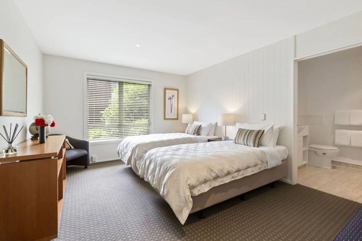 Habitación de hotel con dos camas, un escritorio, una silla, una ventana con persianas, un baño y un espejo.