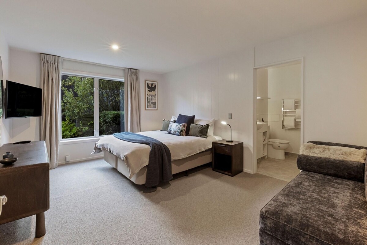 A bedroom with a bed, bedside table, lamp, and a view of greenery outside.