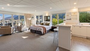 A spacious bedroom with a large bed, a sitting area with two chairs, a desk, and a view of mountains through large windows. - Queenstown House (Queenstown)