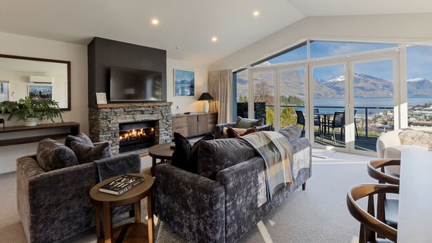 A modern living room with a stone fireplace, comfortable seating, and a large balcony with a scenic mountain view. - Queenstown House (Queenstown)