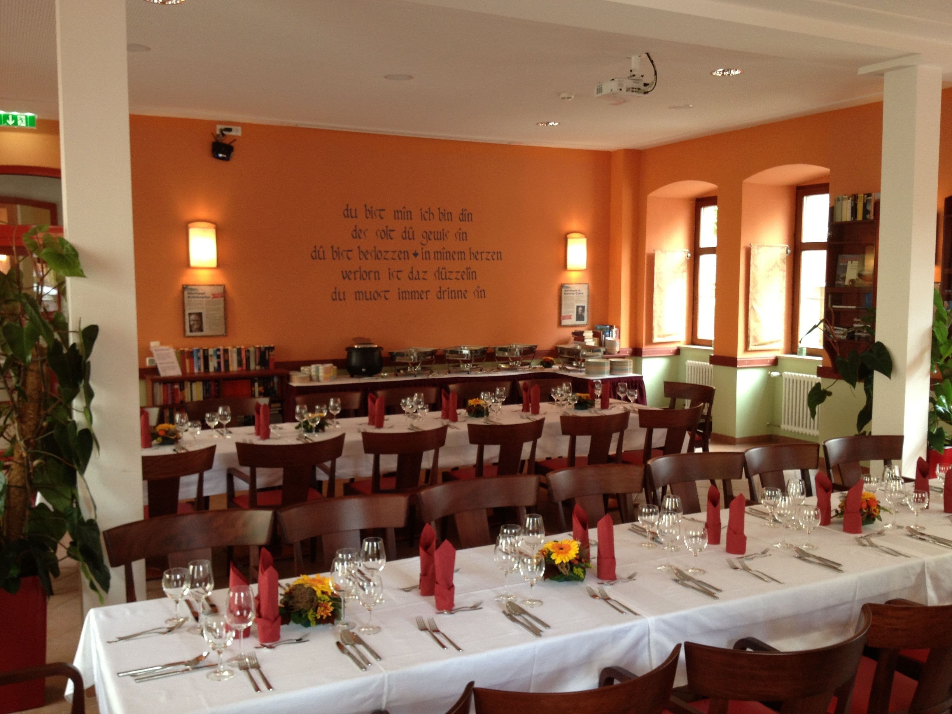 Restaurante com várias mesas preparadas para refeições, apresentando uma parede com texto em língua estrangeira e um projetor no teto.