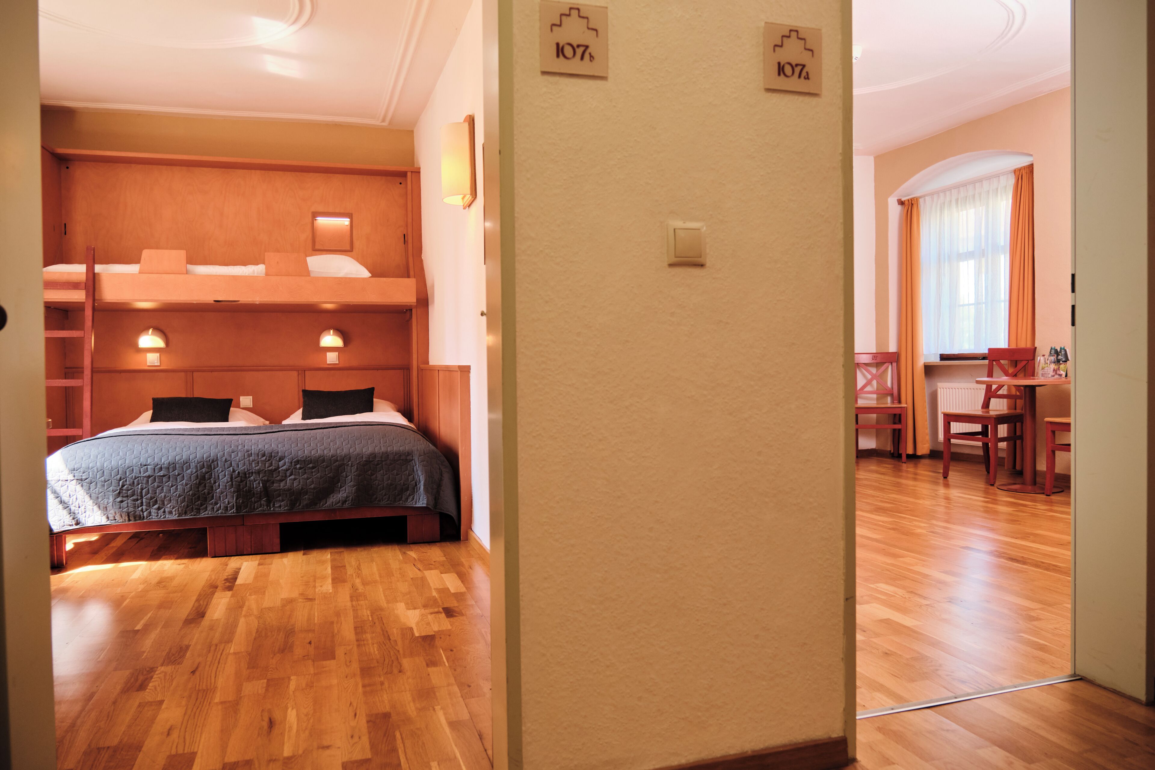 Habitación de hotel con literas, escritorio y sillas. Se ven dos interruptores de luz en la pared y una ventana con cortinas.