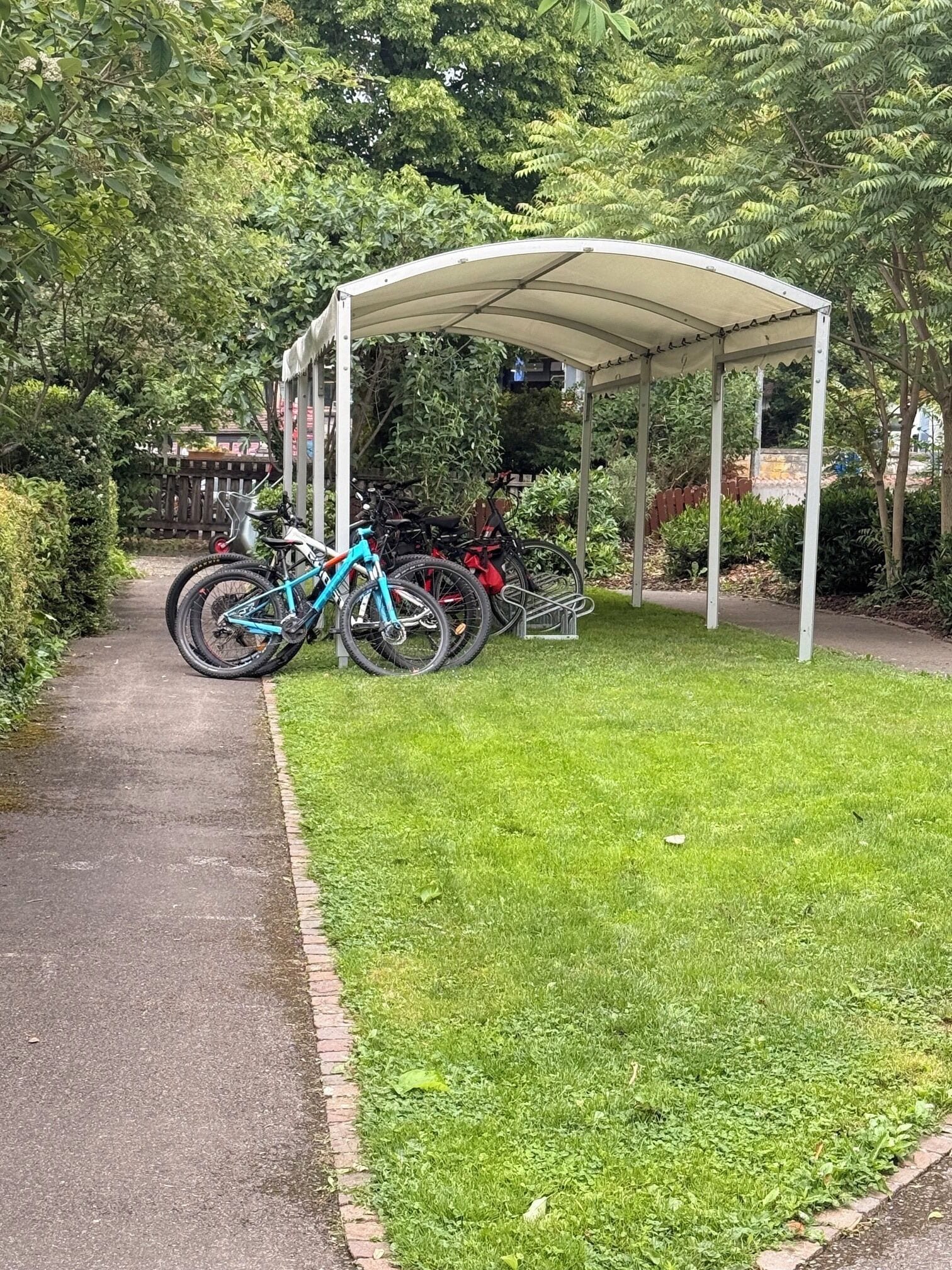 Een overdekt buitengebied met fietsen geparkeerd onder een witte luifel, omgeven door groen.