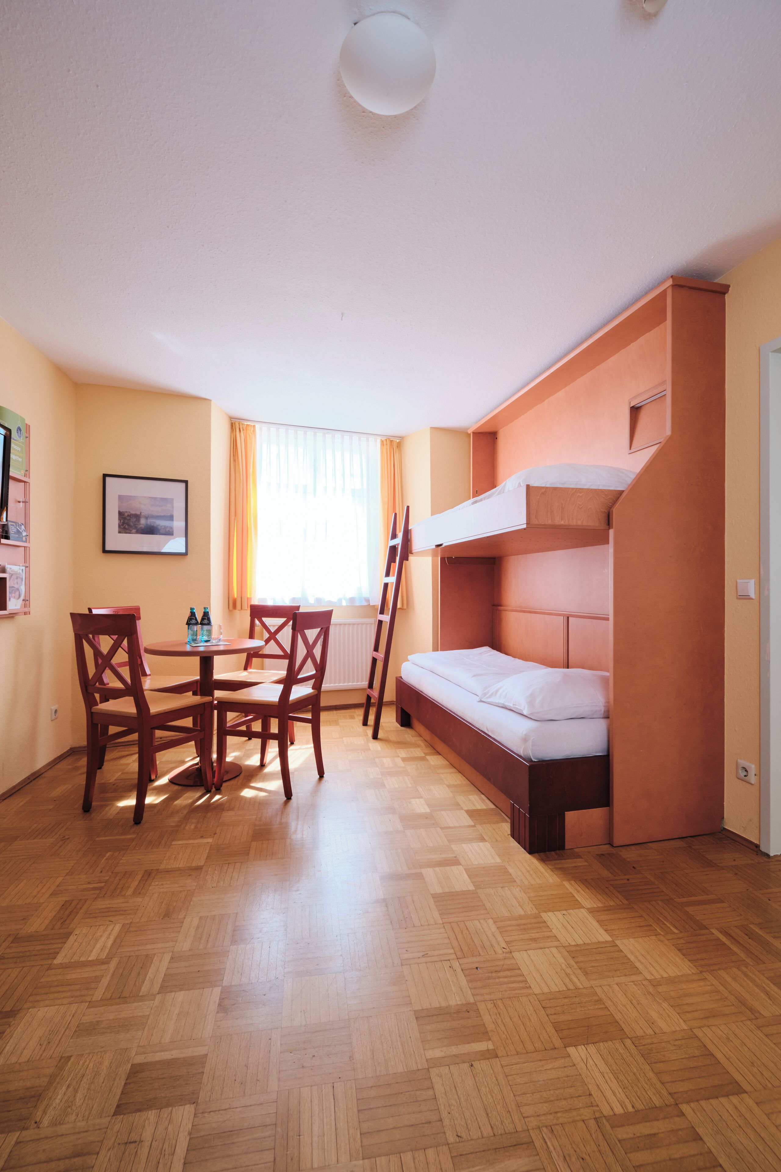 Una habitación con una litera, suelo de madera, una mesa de comedor con sillas y una ventana con cortinas.