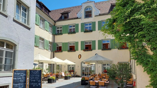Un patio con mesas al aire libre, un menú en una pizarra y un edificio con contraventanas verdes.