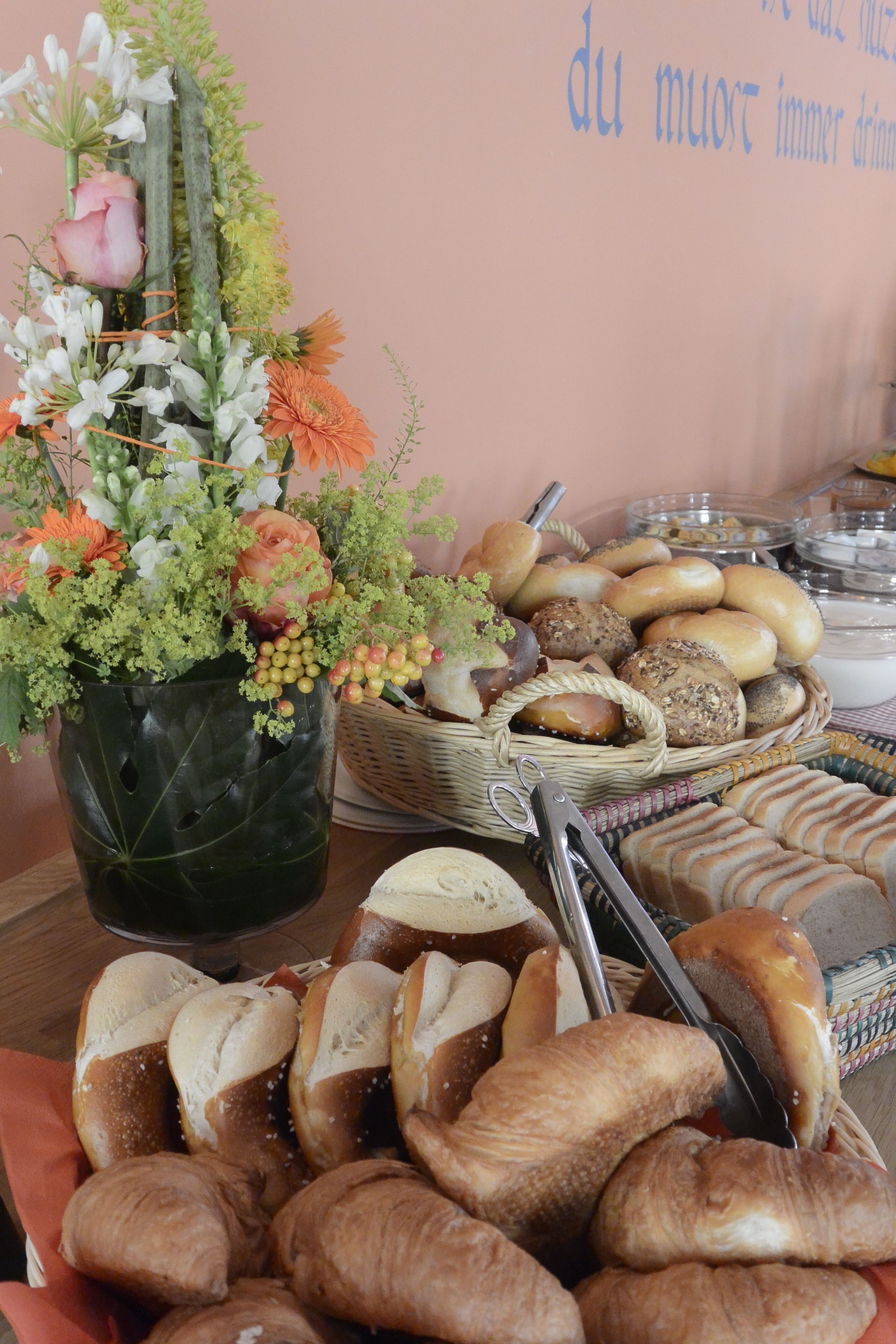 A buffet table with various breads and pastries, a floral arrangement, and a basket of rolls.