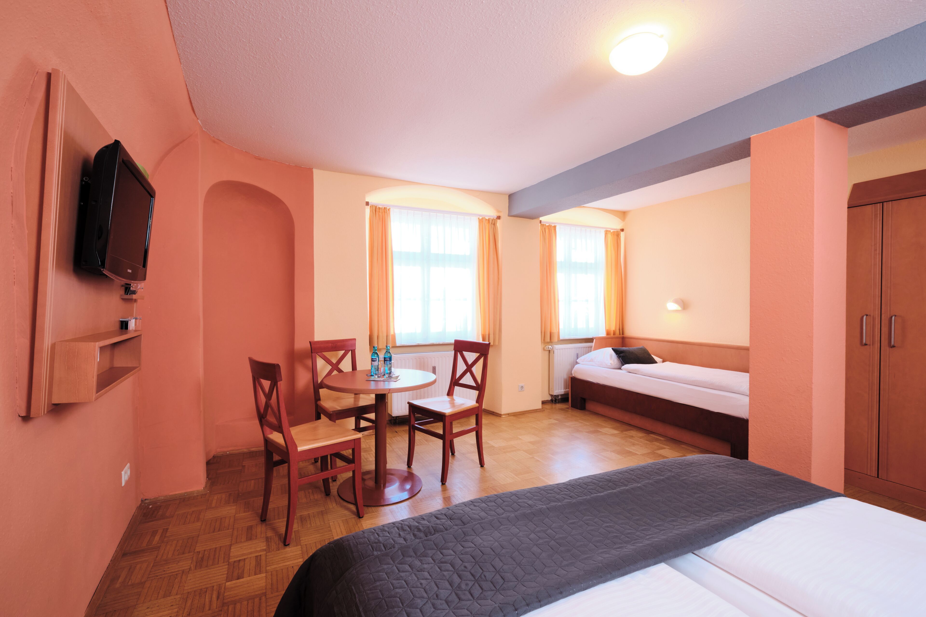 A hotel room with a bed, a desk with chairs, a television mounted on the wall, and a window with curtains.