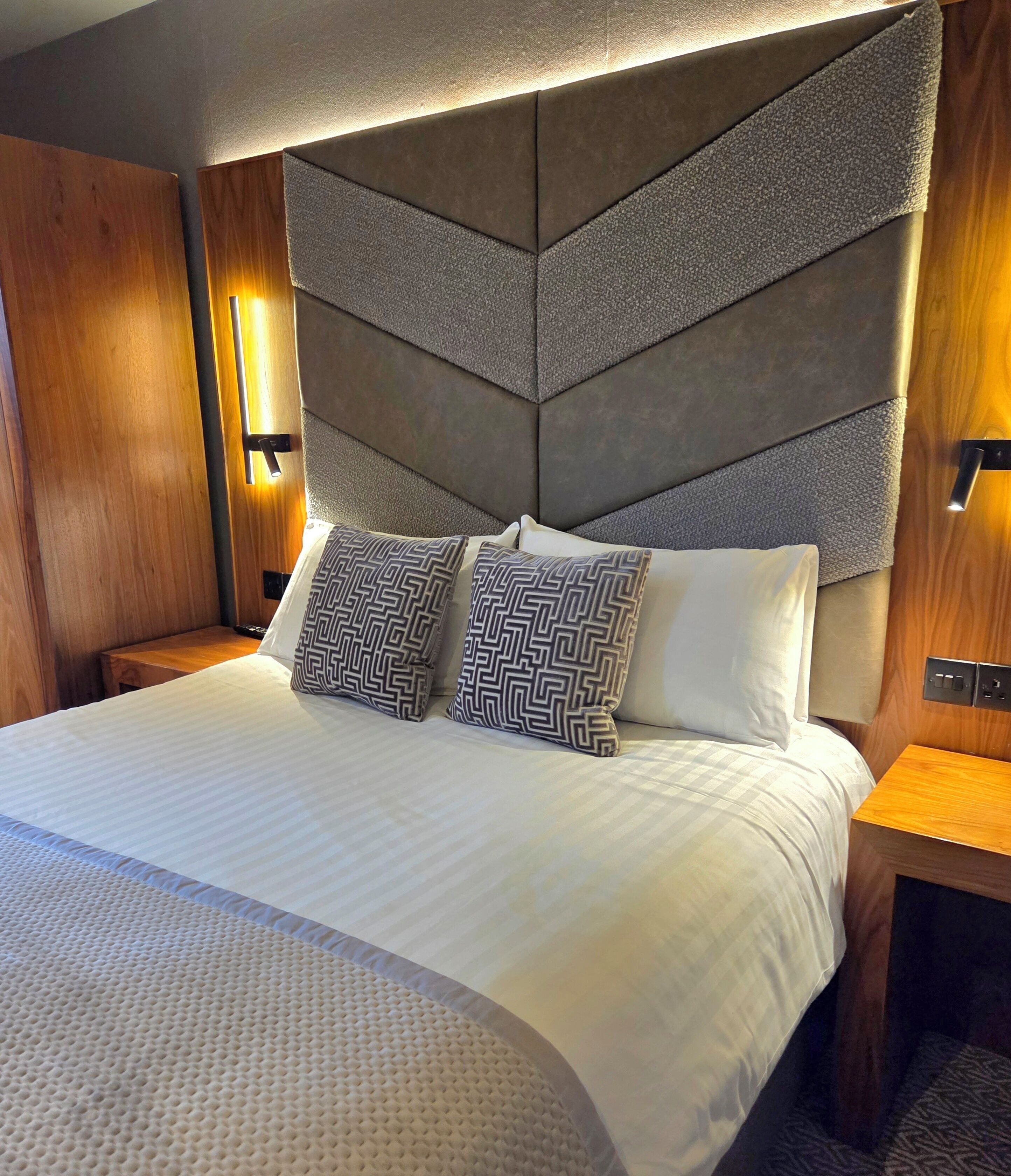 A modern hotel room with a large bed, geometric headboard, and wooden accents.