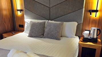 A hotel room with a large bed, a headboard with a geometric pattern, two wall-mounted lamps, a bedside table with a coffee maker, and a rolled towel on the bed.