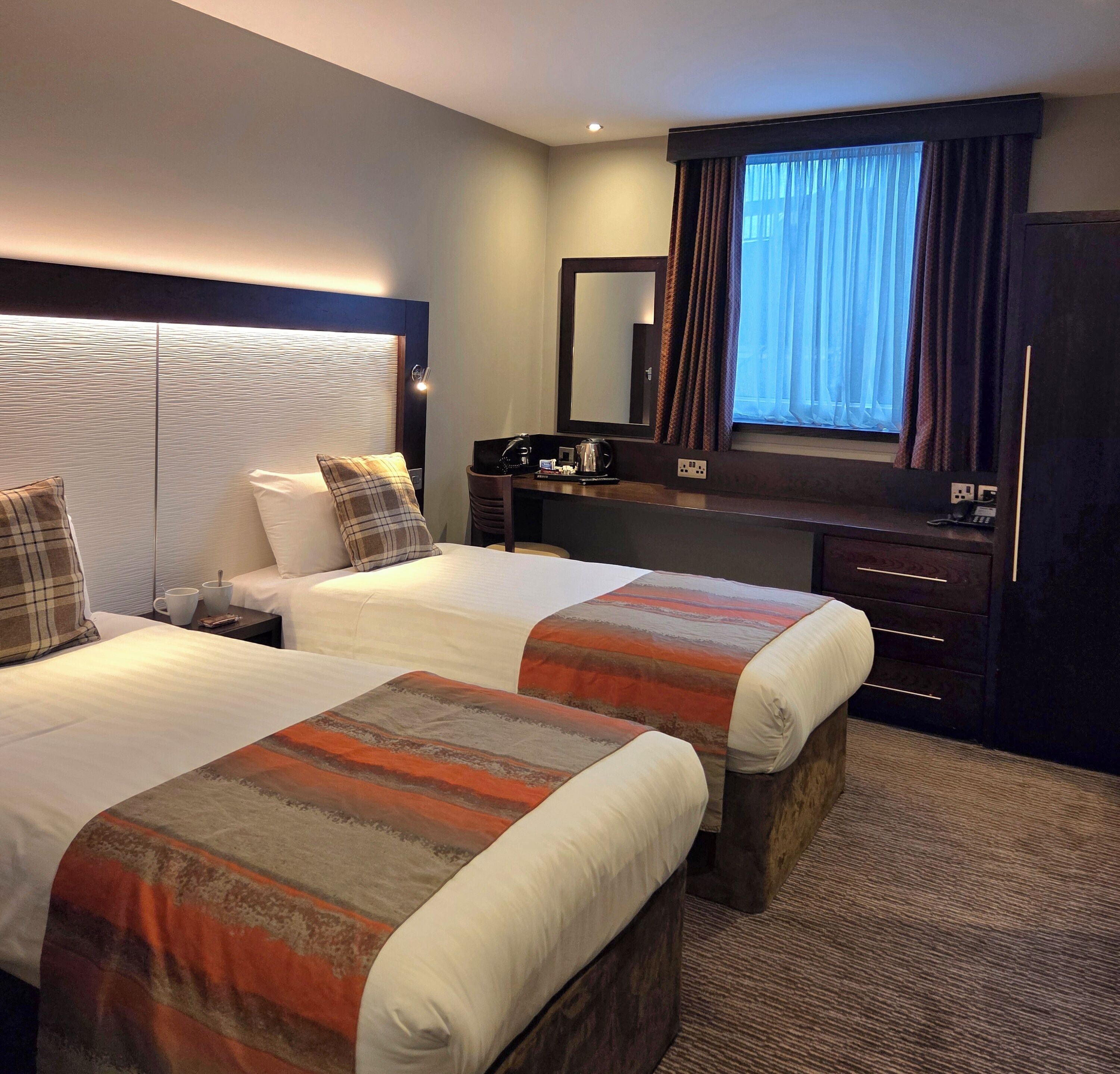 Habitación de hotel con dos camas, un escritorio, una silla, un espejo y una ventana con cortinas.