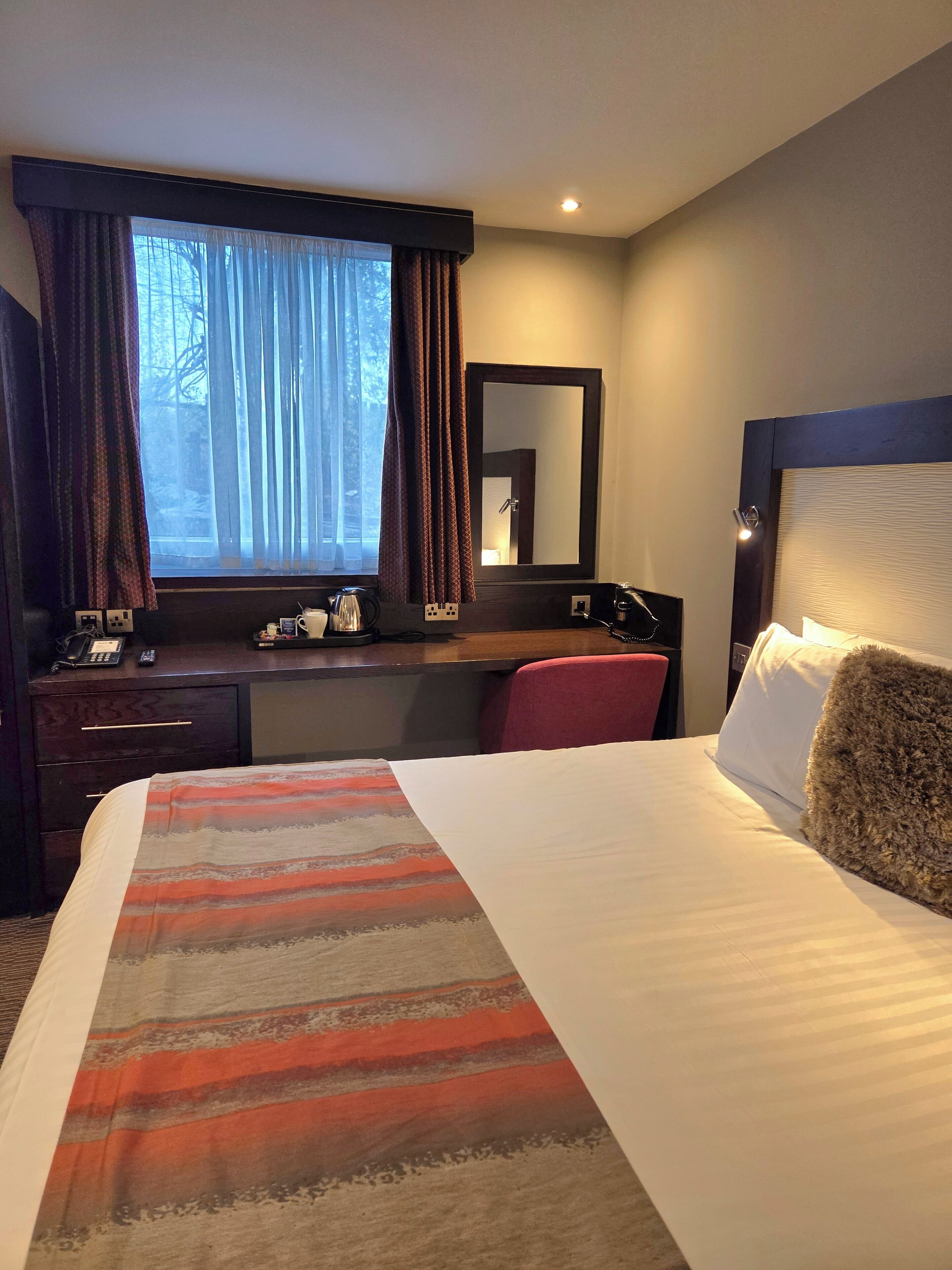 A hotel room with a bed, a desk, a chair, a window with curtains, a mirror, and a lamp.