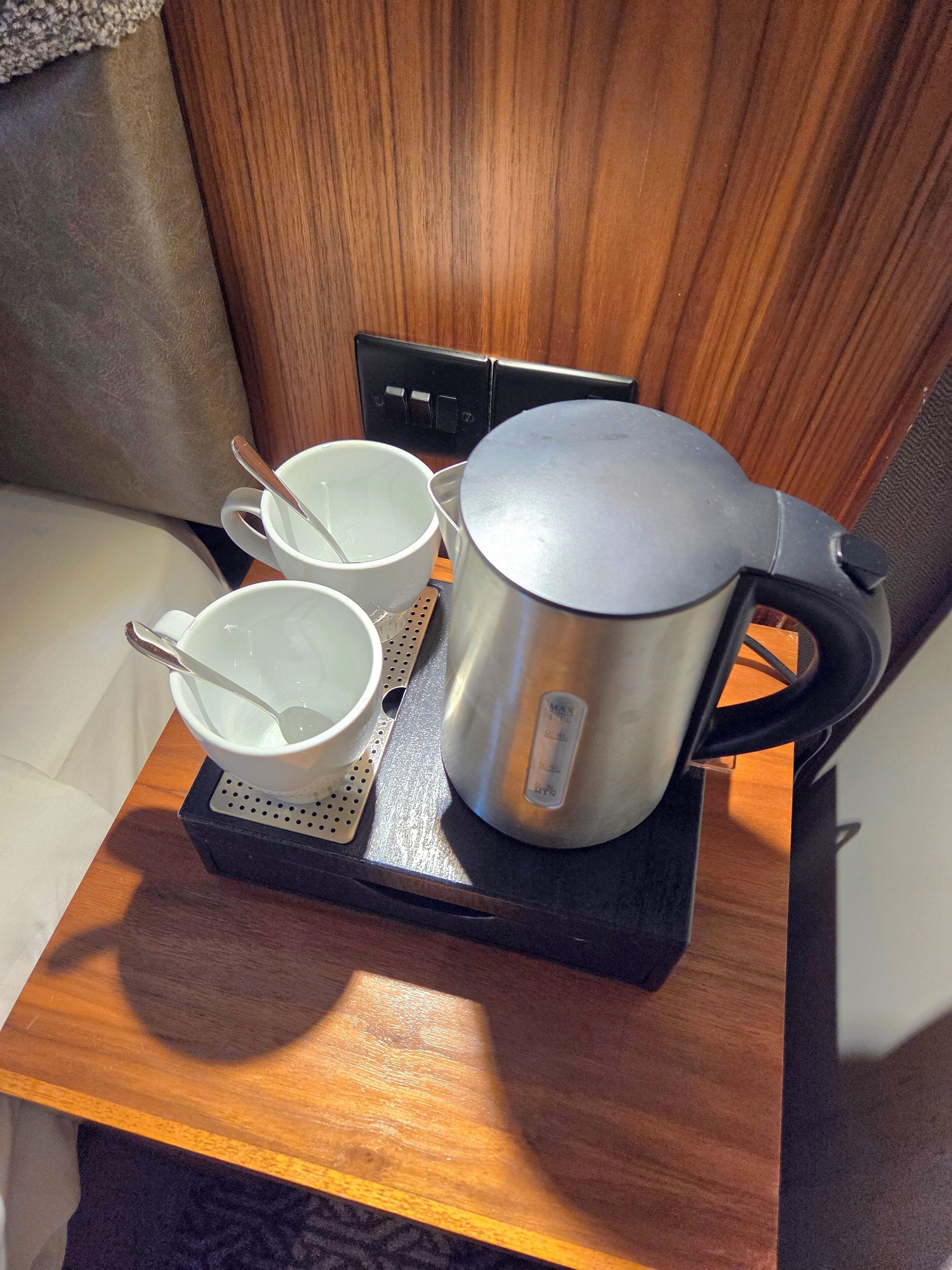 A kitchen area with a stainless steel kettle, two white cups with spoons, and a black tray on a wooden countertop.