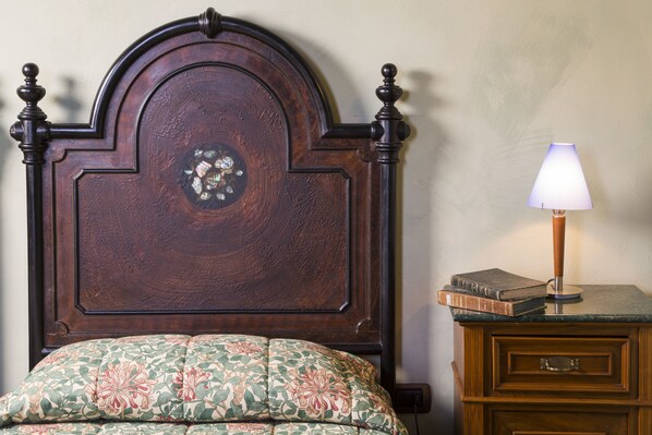 Una cama de madera con un cojín estampado con motivos florales, una mesita de noche con una lámpara y un libro.