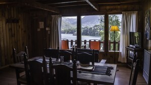 A dining area with wooden furniture, a table with cutlery, a television, and a view of a lake and mountains through large windows. - Cabañas Villa Pañil (San Carlos de Bariloche)
