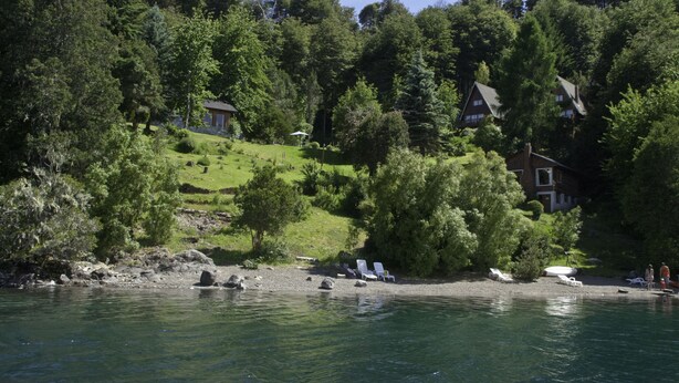 A lakeside property with a house surrounded by dense forest, a grassy area, and a dock with a boat. - Cabañas Villa Pañil (San Carlos de Bariloche)