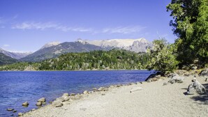 A clear view of a lake with mountains in the background, a rocky shoreline, and some trees. - Cabañas Villa Pañil (San Carlos de Bariloche)