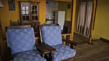 Una habitación con dos sillones azulados estampados, un aparador de madera y una cocina al fondo.