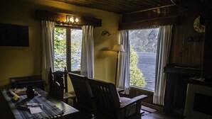 A cozy room with a wooden table, chairs, a lamp, and a view of a lake through large windows. - Cabañas Villa Pañil (San Carlos de Bariloche)