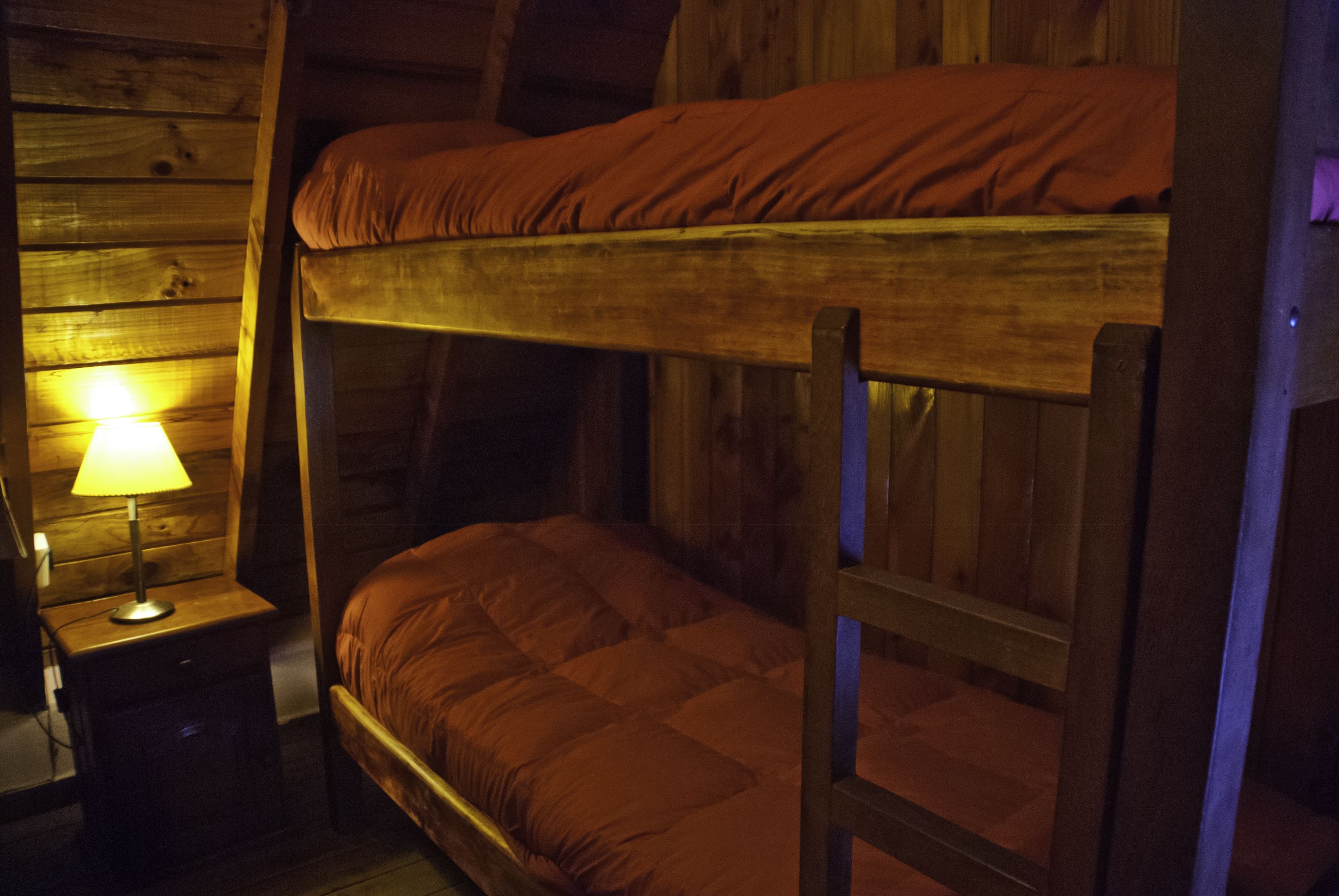 Un letto a castello con struttura in legno e materasso rosso. Accanto al letto, un comodino con una lampada.