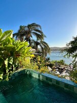 A poolside area with a view of the sea, surrounded by lush greenery and palm trees.